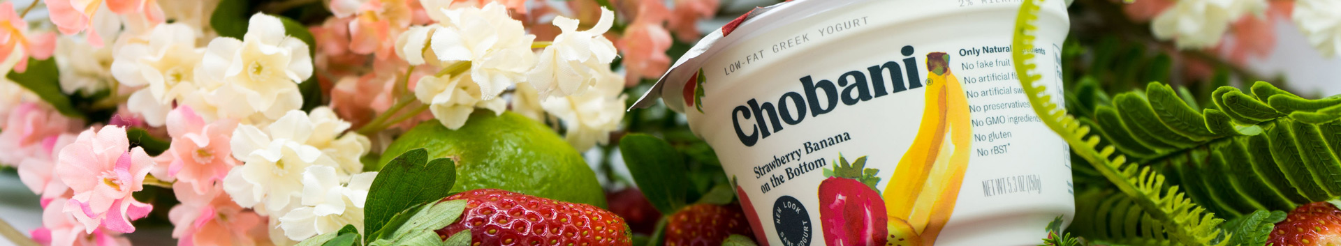 A cup of strawberry banana Chobani yogurt sitting in a bed of fruit, flowers and shrubbery.